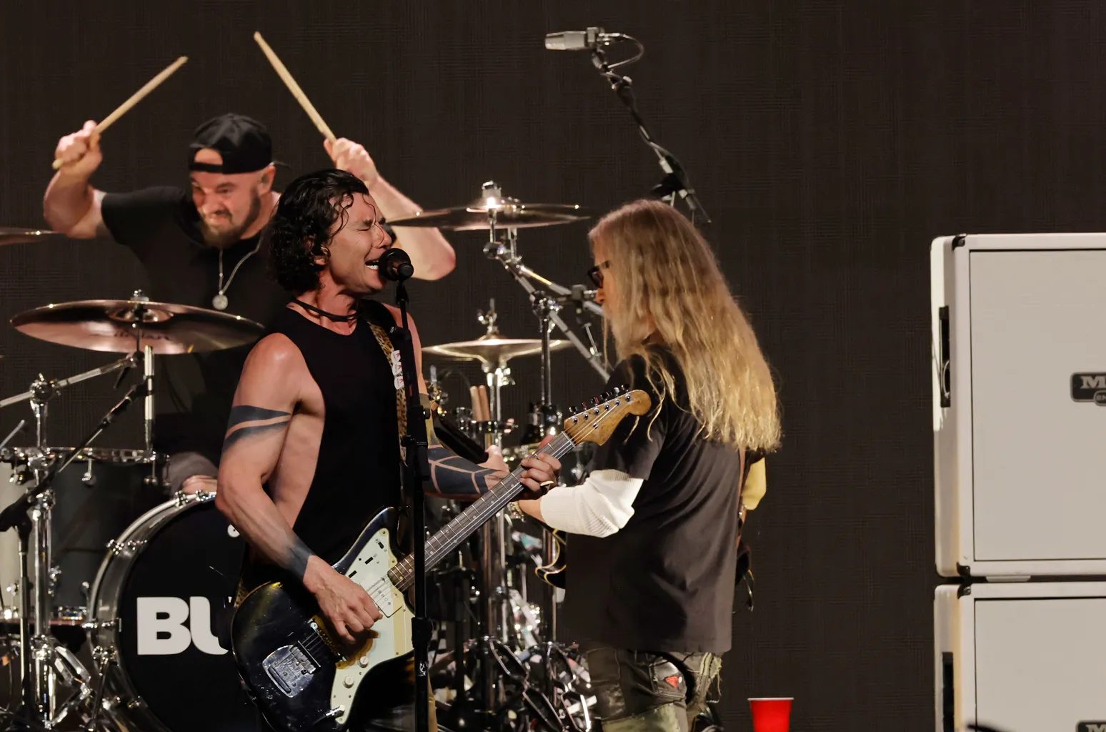 Band performing on stage: a singer with a guitar, a guitarist with long hair and a drummer raising drumsticks